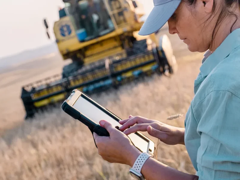 Producer checking GrainX Command on a tablet in the field with a combine harvesting behind them
